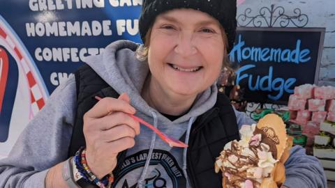 Nicola Webb in a grey hooded top and black woolly hat enjoys a pudding