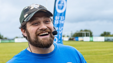A man standing on a grassy field smiles broadly. He wears a blue T-shirt, a camouflage baseball cap, and a headset microphone. Behind him, a blue vertical banner waves in the breeze.