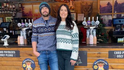 Abbey Matthews and her partner Kyle wearing Nordic style jumpers standing in front of one of their Bar At Yours stalls- they both have jeans on and are smiling in front of bar pumps. 