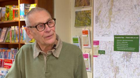 A man with short grey hair and brown glasses, wearing a green top with beige collar, stood in front of a map