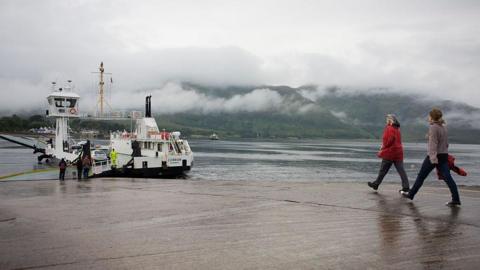 The Corran Ferry is a white and back boat. It is berthed at a slipway. Two women walk down the slipway towards the ferry. Mist hangs on the hills on the opposite shore.