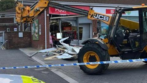 A yellow JCB digger in front of shop with a smashed up window and police tape around the scene 