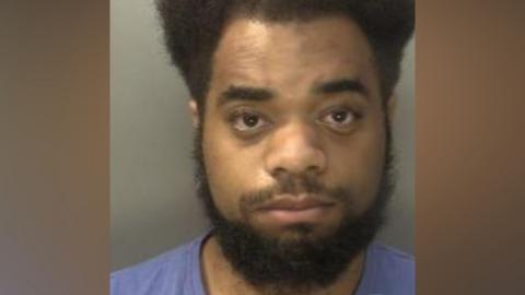 A man who has dark hair curly hair and a dark beard is photographed in front of a neutral background. He has a light blue t-shirt on and appears expressionless.