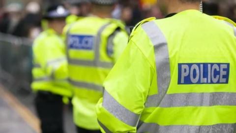The back of a police officer who is wearing a yellow hi-vis jacket with silver reflective strips. It says "police" on the back. In the background are two other officers, wearing the same jackets as well as black police hats.