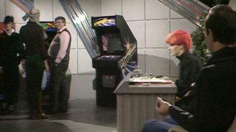 Toyah Wilcox sits behind a desk. A group of men stand around some arcade machines