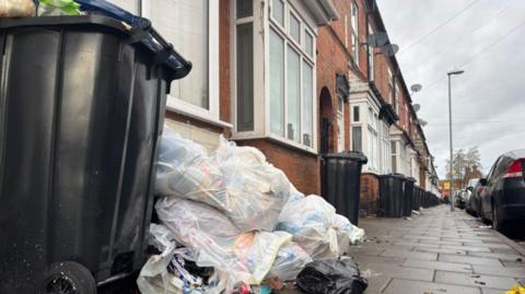 Rubbish is piled up in bags next to a wheelie bin, whose lid is partially open with bags visible in that bin.