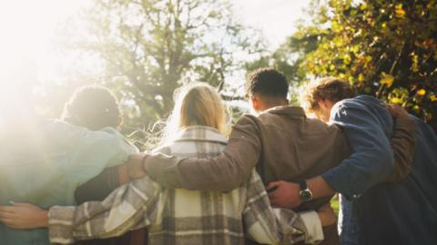 A generic picture of young people, both young men and women, stood linking their arms with their backs to the camera. The sun is shining on them from the top left of the picture.