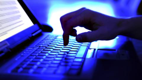 A person's hand is seen typing on a laptop keyboard in a dark room in a stock image. 