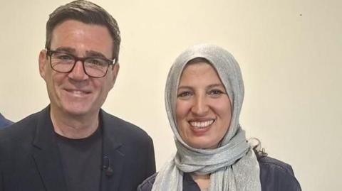 A man in a dark suit and T shirt, with dark-framed glasses, stands with a woman wearing a silver headscarf.