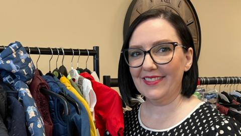 Karen Brown is standing in front of a rack of baby and toddler clothes. She wears black glasses and has bobbed hair. There's a clock on the wall behind her.