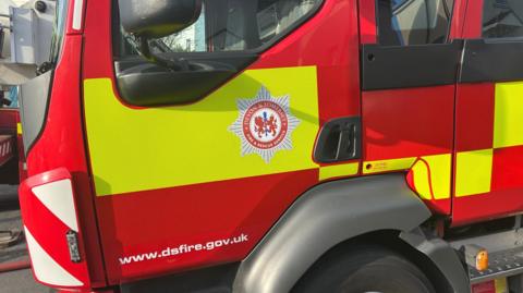 Close-up photo of a Devon and Somerset Fire Service engine. It has red and yellow squares and rectangles. The Devon and Somerset Fire Service badge is on the door of engine.