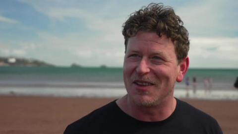John Wells is smiling and looking to the left. He is standing on the beach