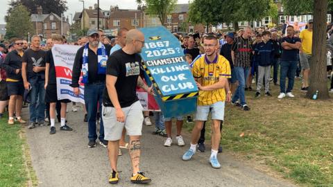 A large group of football fans are walking through a park. Some at the front of wearing football shirts in black and yellow and are carrying a mock coffin painted blue which says "Chansiri killing our club"