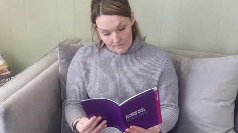 A woman wearing a grey jumper and sitting on a grey sofa. She has brown hair tied back in a ponytail. She is reading a purple booklet bearing the title 'Staying safe, finding hope'.