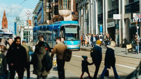 A tram moves along in Birmingham city centre with pedestrians walking nearby. 