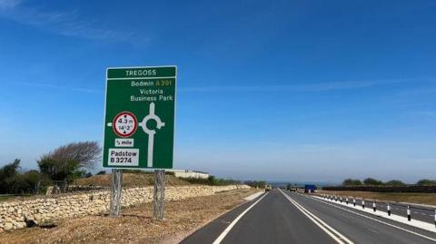 A picture from the side of the road. There is a green sign which shows a roundabout ahead and the arrow pointing up says 'Bodmin and Victoria Business Park" at the bottom of the sign it says "Padstow B3274". The side of the road there is a small stone wall and dirt and grass.