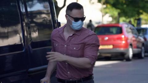 Antony James wearing sunglasses, a blue surgical style face mask and a maroon shirt looks to his left as he approaches a black van.