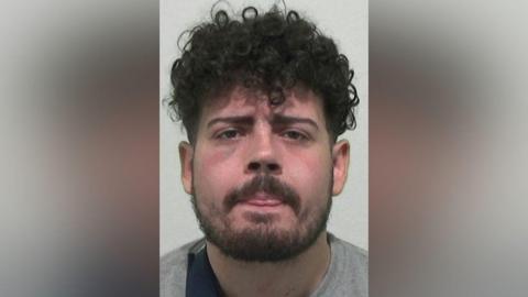Mugshot of Kier Adam-Nuhoglo. He has thick black curly hair, a black beard and a reddened face. He is sticking his tongue out slightly between almost closed lips.