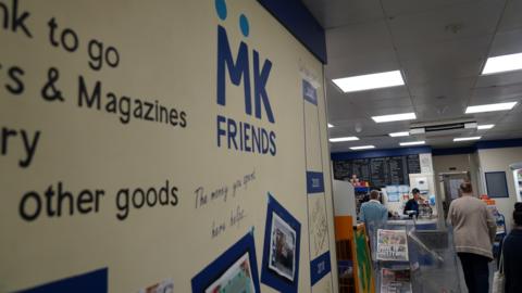 In the foreground is a noticeboard or advertising board, with "MK FRIENDS" branding in blue font printed on it. In the background, there are some people queuing at a counter in the MK Friends shop. Behind the counter on the wall is a menu or price list chalked on a black board.