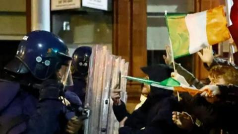 two riot police officers, wearing helmets, standing behind shields facing down a group of young men holding Irish flags