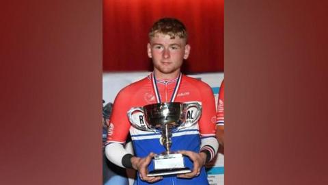 Aidan Worden - a young man with short, blondish hair and a medal around his neck, holds a trophy. He wears a red, white and blue cycling jersey.