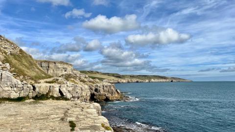 Cliffs on the left and in the distance, the sea on the right, under a blue sky with clouds