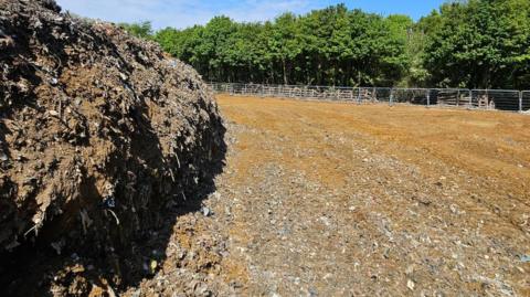 A pile of waste is to the left, but the rest of the picture shows a flat field of soil and dirt, with fencing on the outskirts.