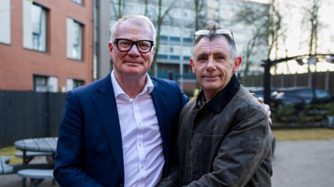 Mayor Richard Parker in a navy suit jacket and white shirt pictured alongside a man in a grey jacket in a carpark outside a building on a grey day. Richard Parker has white hair and thick black glasses. The man on the right has receding greying dark hair and glasses pushed back on his head.