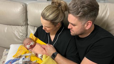 Daniel and Bryony Russo holding his daugher Emily Russo. She is lying on a yellow padded blanket and has a yellow wool blanket over her. Daniel is holding his daugher's hand and looking down at her with sad eyes. He is wearing a black t-shirt and a black watch.