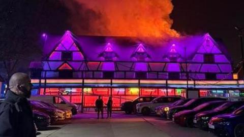 A large Tudor looking building at night time, which is lit in purple neon lights, but there is an orange glow from fire, and smoke tinged with orange billows above it. In the foreground are parked cars and a side-on of a man.