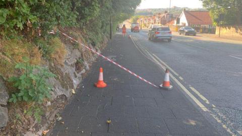 A pavement and road in West Road in Newcastle. There are two cones with some orange tape stretching across it. 