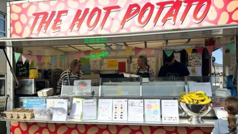 The image shows a food truck called THE HOT POTATO, decorated in red and yellow with potato-themed graphics. Inside the truck are three people and one customer in front. The counter displays a bowl of bananas, printed materials, and other food items. 