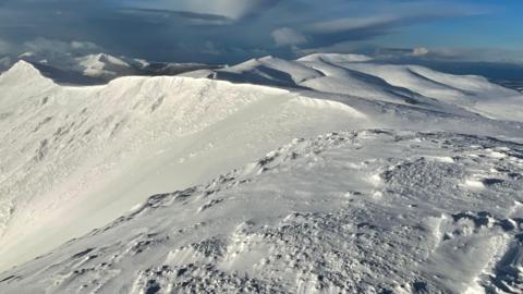 Summit of Blencathra in the Lake District. A snow-topped summit with blue sky and dark clouds in the distance. 