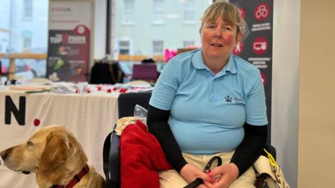 Jenny sits in a chair looking at the camera. She has a light blue polo t-shirt on with a Guide Dogs logo on it with a black underlay and cream coloured trousers. She has a lead in her hands and her Labrador Retriever sitting to her side which is looking to the left of the photo.