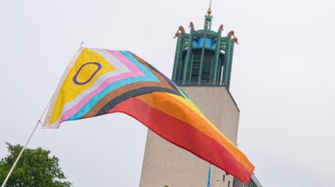 The New Progress Pride Flag being flown outside Newcastle Civic Centre. The flag contains a number of colourful arrows leading into horizontal stripes, which represents a broad spectrum of the LGBTQ+ community. The green, copper tower of the civic hall can be seen in the background, which has several bronze coloured seahorse statues at the top.
