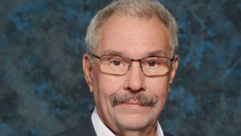 Colin has greying short hair and half-rim glasses and a moustache. He is in front of a blue/black design background and the top of his shirt collar can be seen.