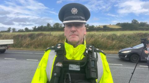 Ch Supt Norman Haslett is standing on the side of a road. He is wearing a PSNI cap and a yellow high viz jacket with a body vest. There's a walkie-talkie attacked to his vest. He has medals on his lapels. Fields are in the background. Some cars are passing. A tripod with a camera attached is to his side. 