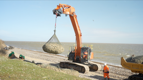 A large piece of machinery loads sea rock bags on to a coastline. Workers in orange hi-vis walk around the beach nearby.