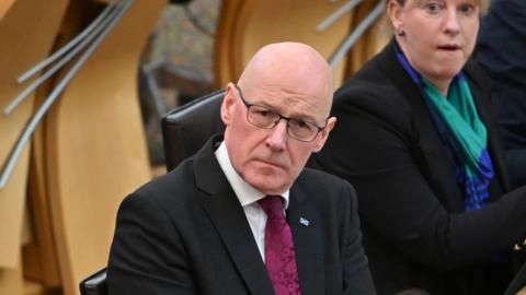 John Swinney in dark suit with a white shirt and purple tie sitting in the Holyrood chamber