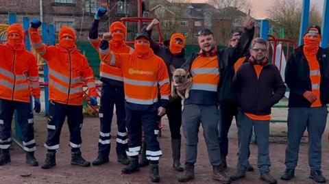 A group of men wearing high-vis orange workwear and orange balaclavas stand with their fists raised in the air on a playground. 