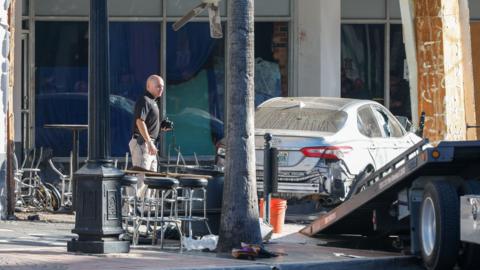 Image of an official standing beside a wrecked car, next to a tow truck, in front of a bar with overturned bar stools