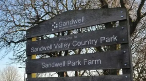 A mainly dark sign that includes the words Welcome to Sandwell Valley Country Park. Trees are in the background.