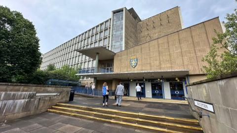 People walk into County Hall. It is a large eight-storey concrete building with lots of windows and the Durham Council logo on the entrance wall.
