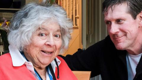A woman with short white hair and a red rugby short grins manically at the camera whilst a man with a dark shirt smiler admiringly at her