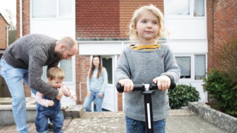 A family are out in front of a house. In the foreground is a young girl with shoulder length blonde hair holding onto the handles of a scooter. In the background a man is helping a toddler to stand and further behind that a women looks towards them.