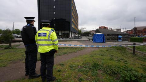 Two police officers stand with their backs to us by blue and white police tap with surrounds paths, grass and paving - with a blue tent on the paving.