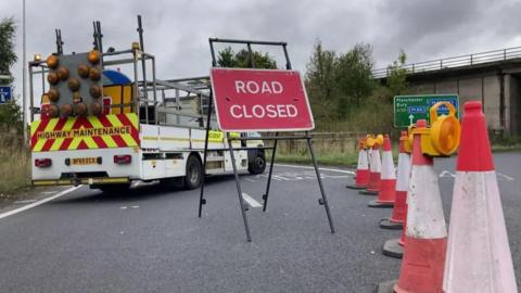 A red "road closed" sign sits behind a row of traffic cones blocking off a slip road to the M65 near Burnley. A highway maintenance van stands behind the sign.