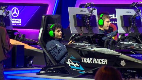 Young man with bright green headphones sitting in gaming chair in front of a screen driving a gaming wheel