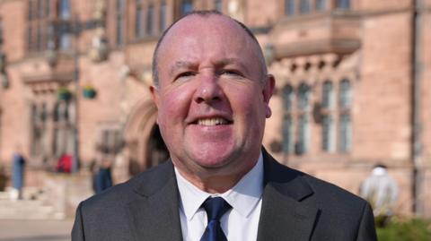Jim O'Boyle smiles at the camera in front of Coventry's Council House. He is wearing a grey suit jacket and a white shirt with a navy blue tie. 