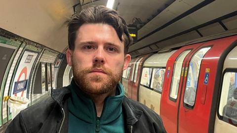 Niall McNamee, standing on a London underground platform while a tube passes on the left-hand side. Niall has dark, short hair, and a beard. He is wearing a black jacket, with a green fleece underneath.